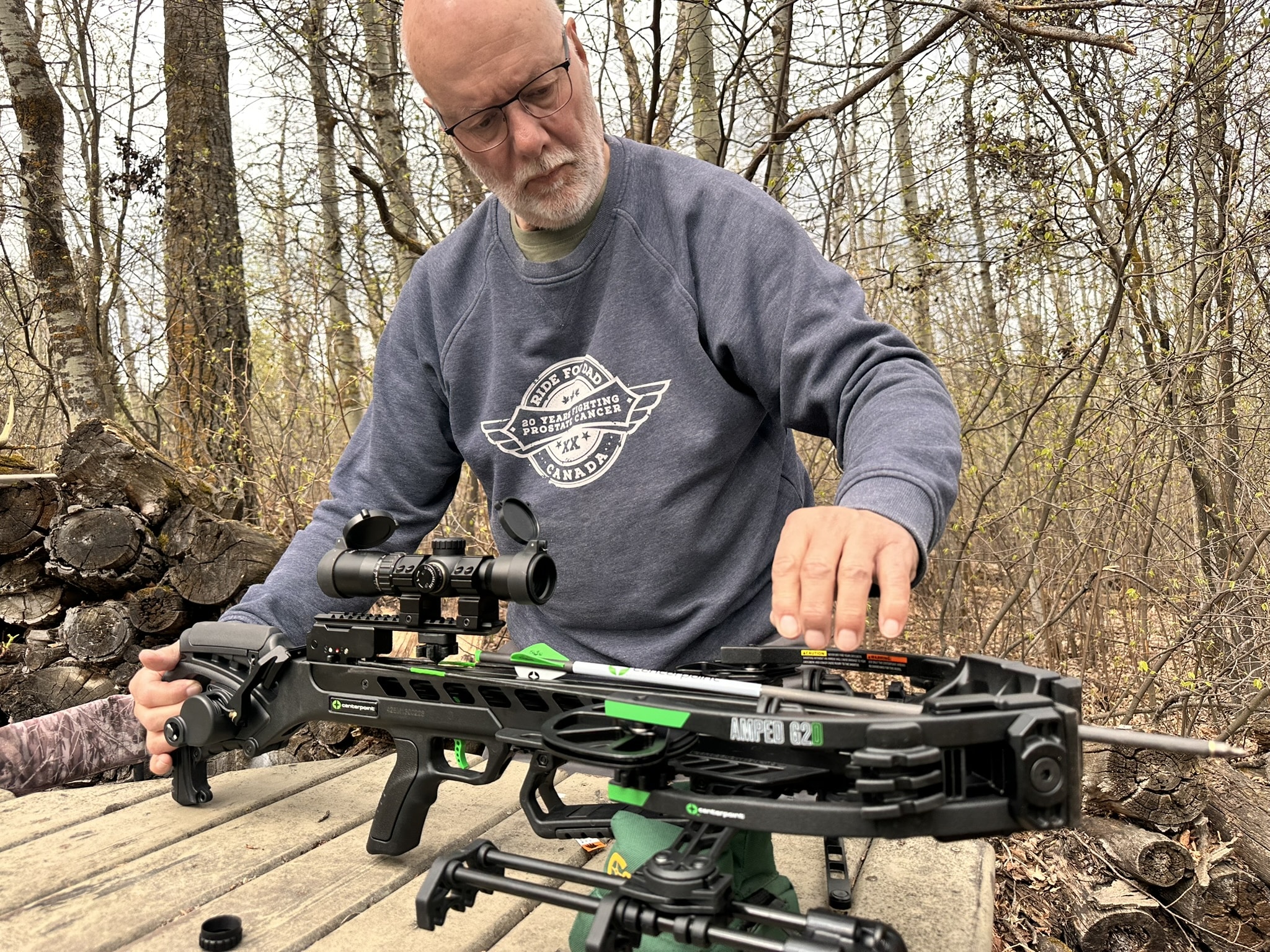 Man with glasses in a gray sweatshirt outdoors, adjusting a black crossbow on a wooden bench in a forest.