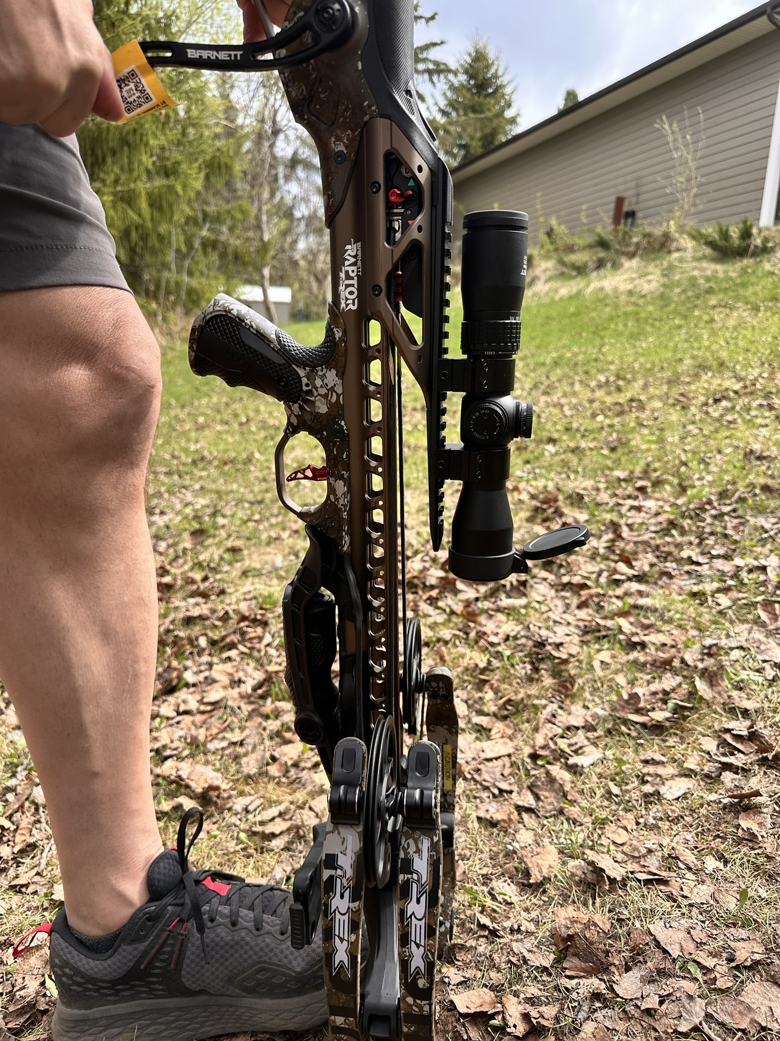 Close-up of a camo compound bow with a mounted scope and stabilizer, held by a person wearing gray sneakers outdoors.