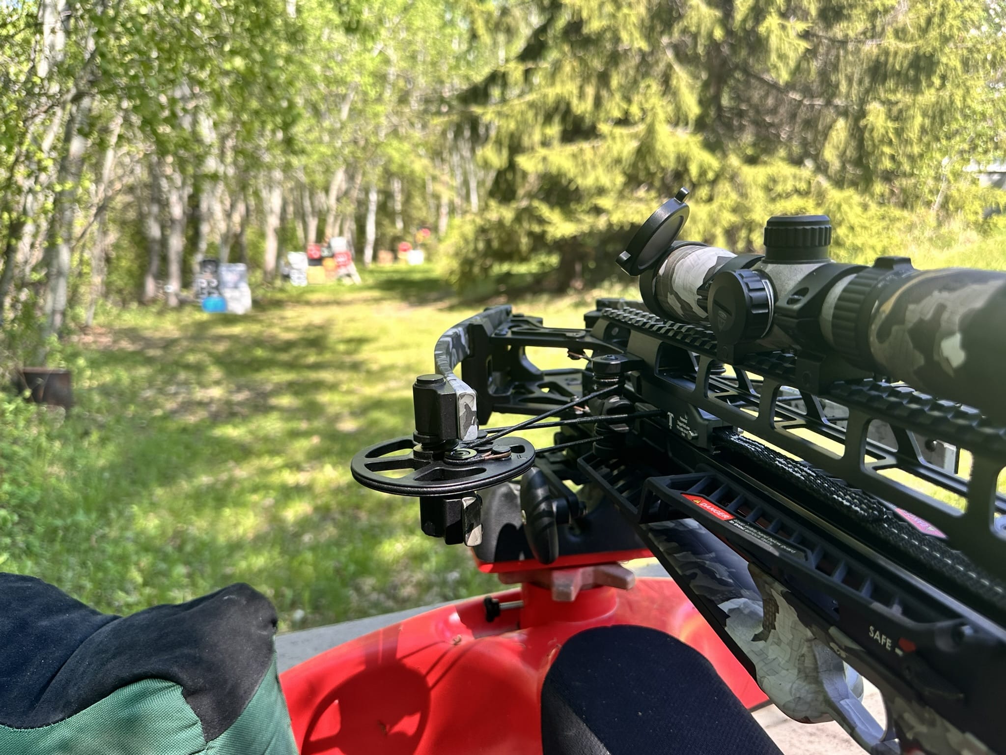 Close-up of a camouflage scope mounted on a crossbow, outdoors in a green, wooded area with trees in the background.