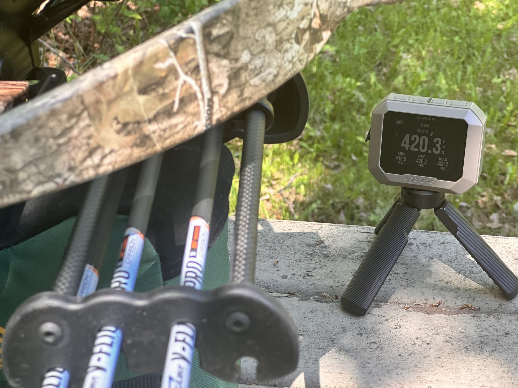 Close-up of a camouflaged hunting setup with a Garmin bow-mounted device and tripod on a wooden surface.