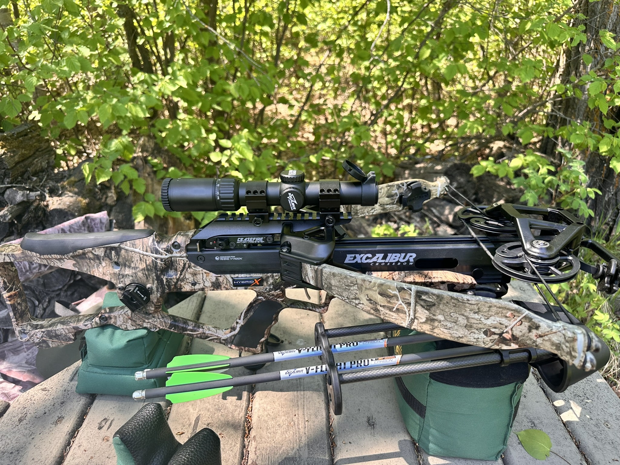 Camouflage crossbow with scope and green-tipped arrows resting on gear among leafy outdoor vegetation.
