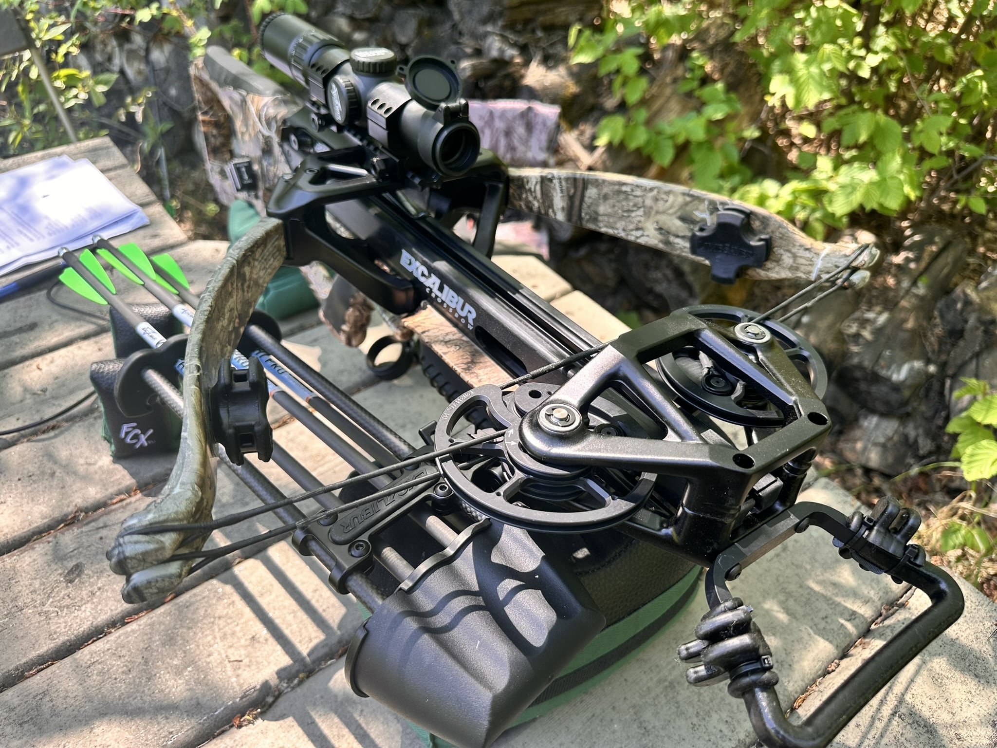 Close-up of an Excalibur hunting crossbow with a mounted scope, resting on a camo frame outdoors.