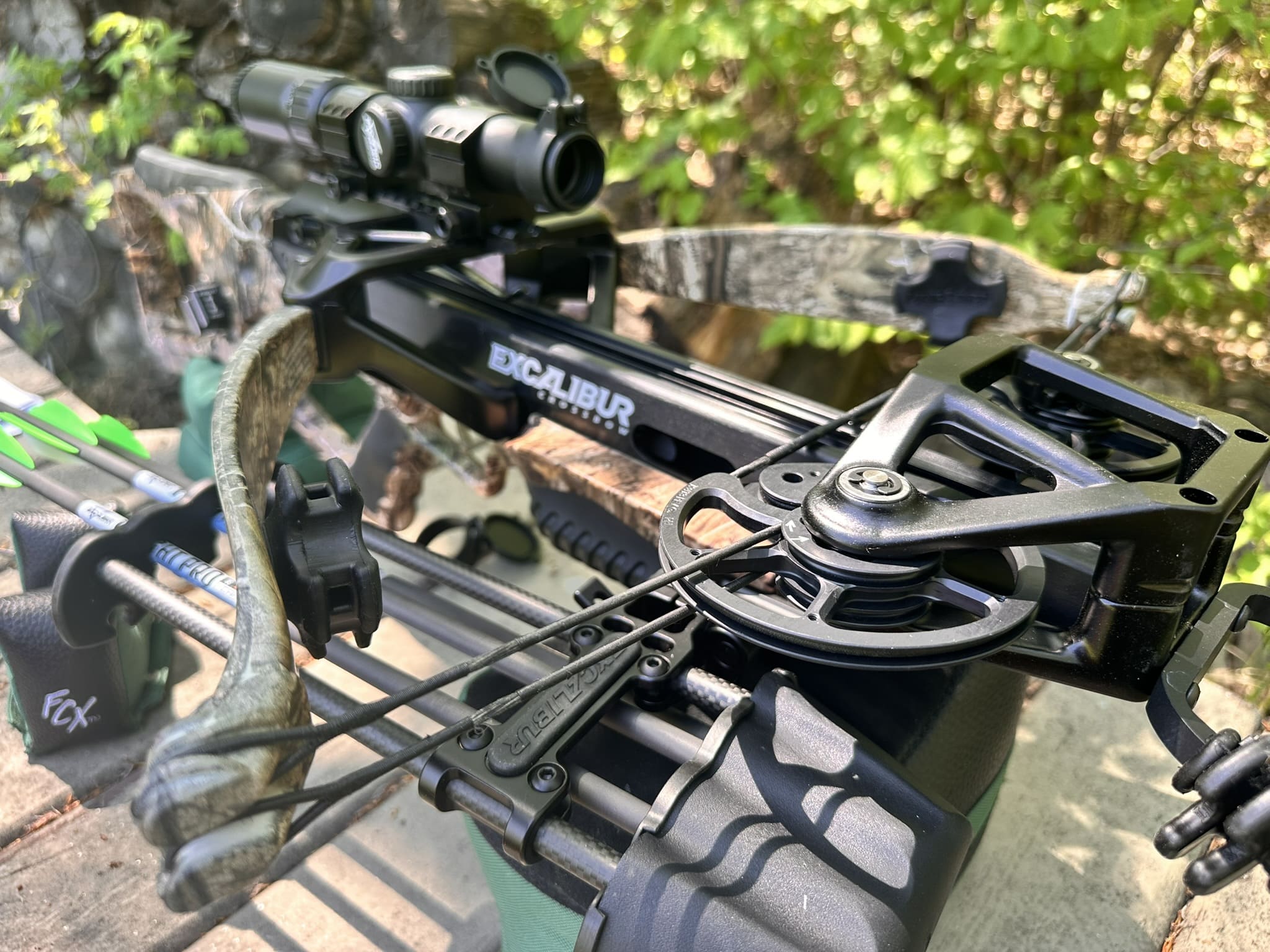 Close-up of a modern crossbow with a scope mounted on top, outdoors among green foliage (Excalibur branding visible).