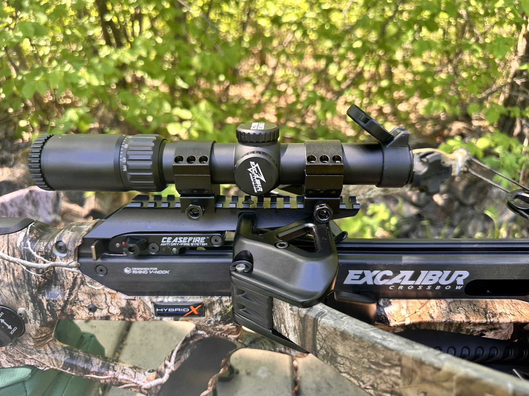 Crossbow with a mounted scope and camo finish, outdoors in a wooded area; close view of the scope and rail system.