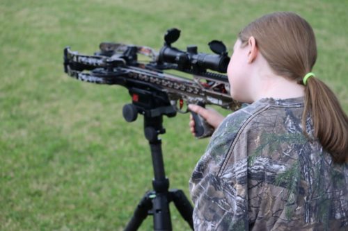 Person in camouflage aiming a modern scoped crossbow on a bipod in a grassy field.