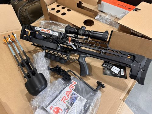 Ravin R470 crossbow with scope and orange-fletched arrows in a cardboard box, surrounded by packing material and accessories.