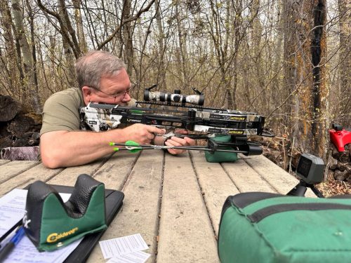 Man lying prone in a forest, aiming a camouflaged crossbow with a scope, surrounded by archery gear and bags.
